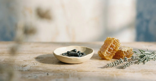 Editorial still-life of a small ceramic dish with botanical supplement capsules beside a raw honeycomb fragment and dried lavender on pale oak — calm, warm, and natural, evoking the body's balanced inflammation response.
