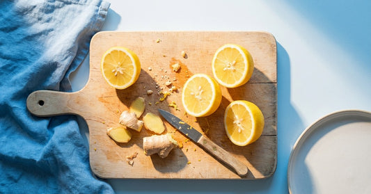 An overhead shot of a wooden chopping board with halved lemons and a peeled ginger root, raking morning light, citrus-yellow and white palette. ENERGY palette.