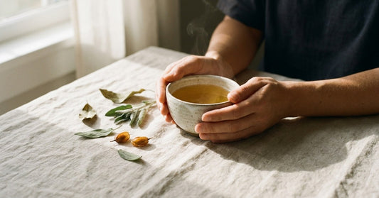 Editorial lifestyle photograph: relaxed hands cupping a small handmade ceramic bowl of warm golden liquid on a raw linen tablecloth with dried botanical leaves nearby, evoking calm considered decision-making.