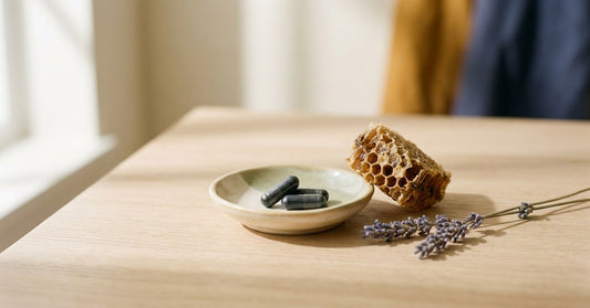Editorial wellness still-life of a handmade ceramic dish with botanical supplement capsules beside a honeycomb fragment and dried lavender on pale oak — evoking quiet inner balance and the rhythm of cells.