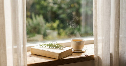 A closed book with a sprig of fresh rosemary resting on its cover beside a steaming ceramic cup of herbal tea on a sun-warmed wooden windowsill with sheer linen curtains and green foliage visible outside