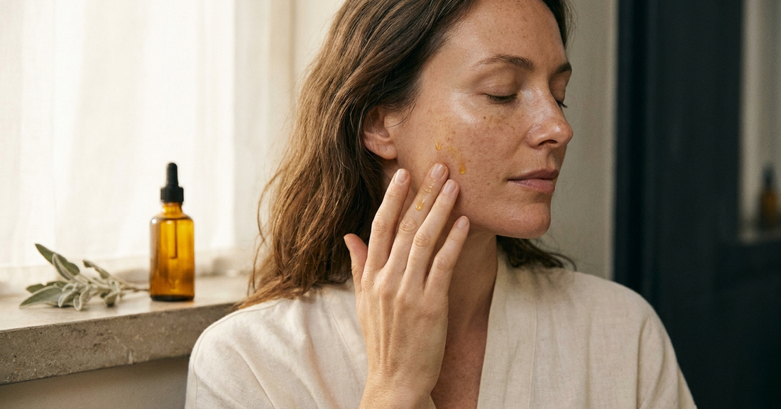 A woman in soft morning light gently pressing a few drops of amber EBC-46 Blushwood Berry tincture into her cheekbone — a quiet, plant-based skin renewal ritual.