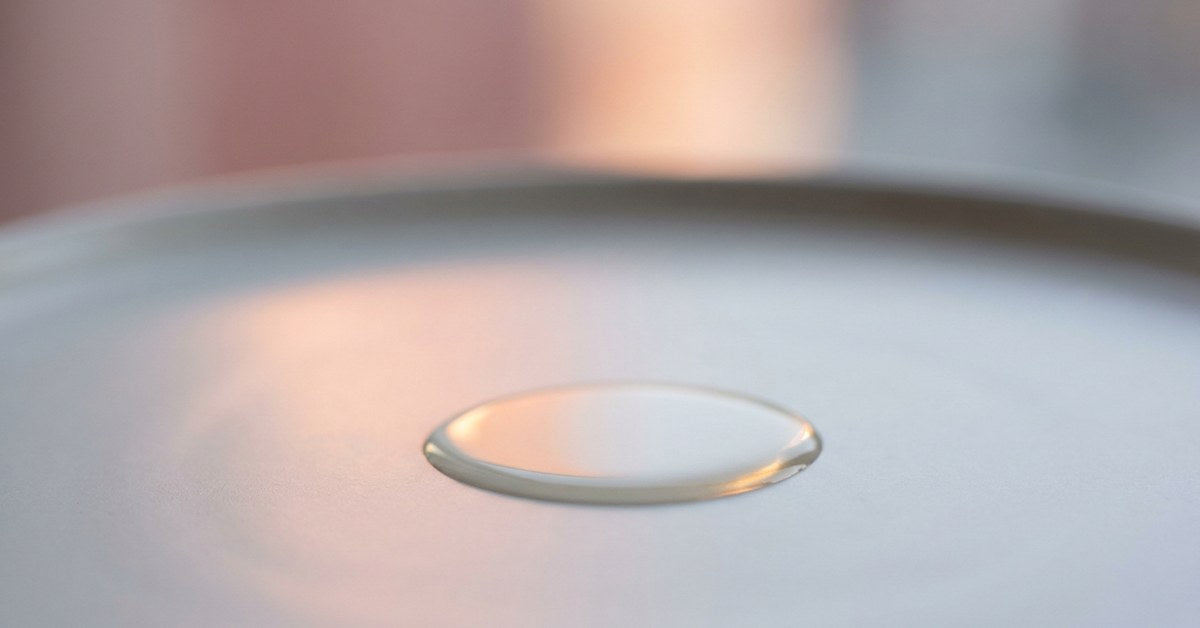 A single drop of clear viscous oil spreading slowly on a smooth pale-grey ceramic surface, peach reflection from a window beyond. SKIN palette.