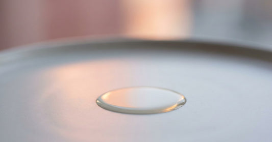 A single drop of clear viscous oil spreading slowly on a smooth pale-grey ceramic surface, peach reflection from a window beyond. SKIN palette.