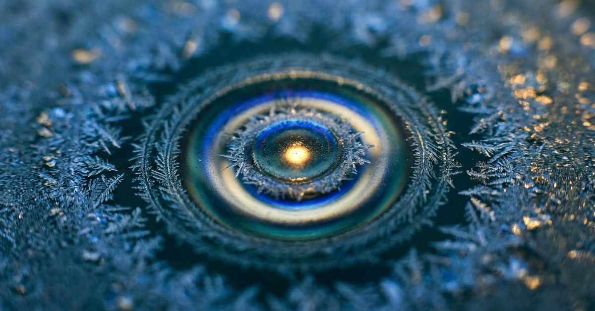 Macro of frost forming concentric crystal patterns on dark glass, single warm pinpoint of refracted light at the centre. IMMUNE palette.